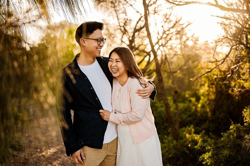 A smiling couple stands close together on a sunlit path surrounded by trees, with the man’s arm around the woman as they both laugh and enjoy the outdoors during a family-photography-williamstown-beach session.