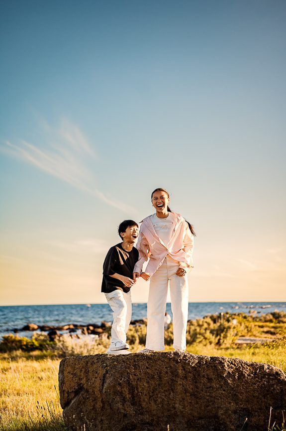 A boy and a girl stand on a large rock in a grassy area near the sea, laughing together under a clear blue sky during sunset—a perfect spot for memorable photography session locations.
