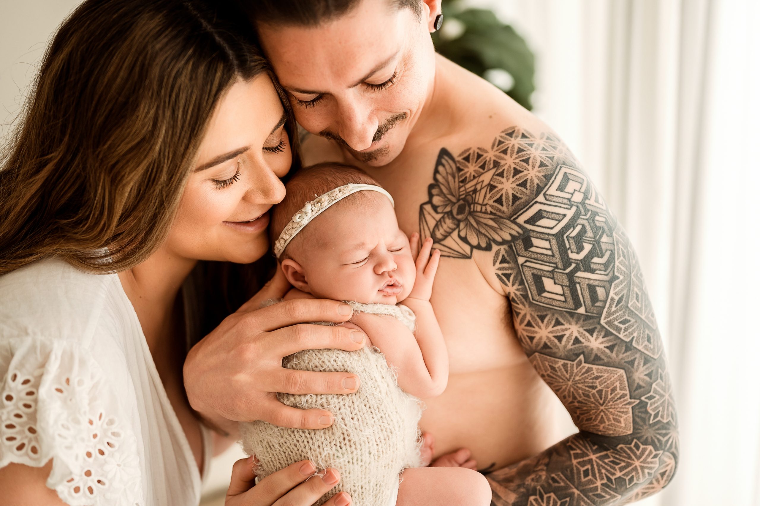 A smiling couple lovingly embraces their sleeping newborn baby. Captured by a Family and Newborn Photographer Melbourne West, the man’s geometric tattoo and the baby’s knitted outfit shine in warm, natural light filling the cozy setting.