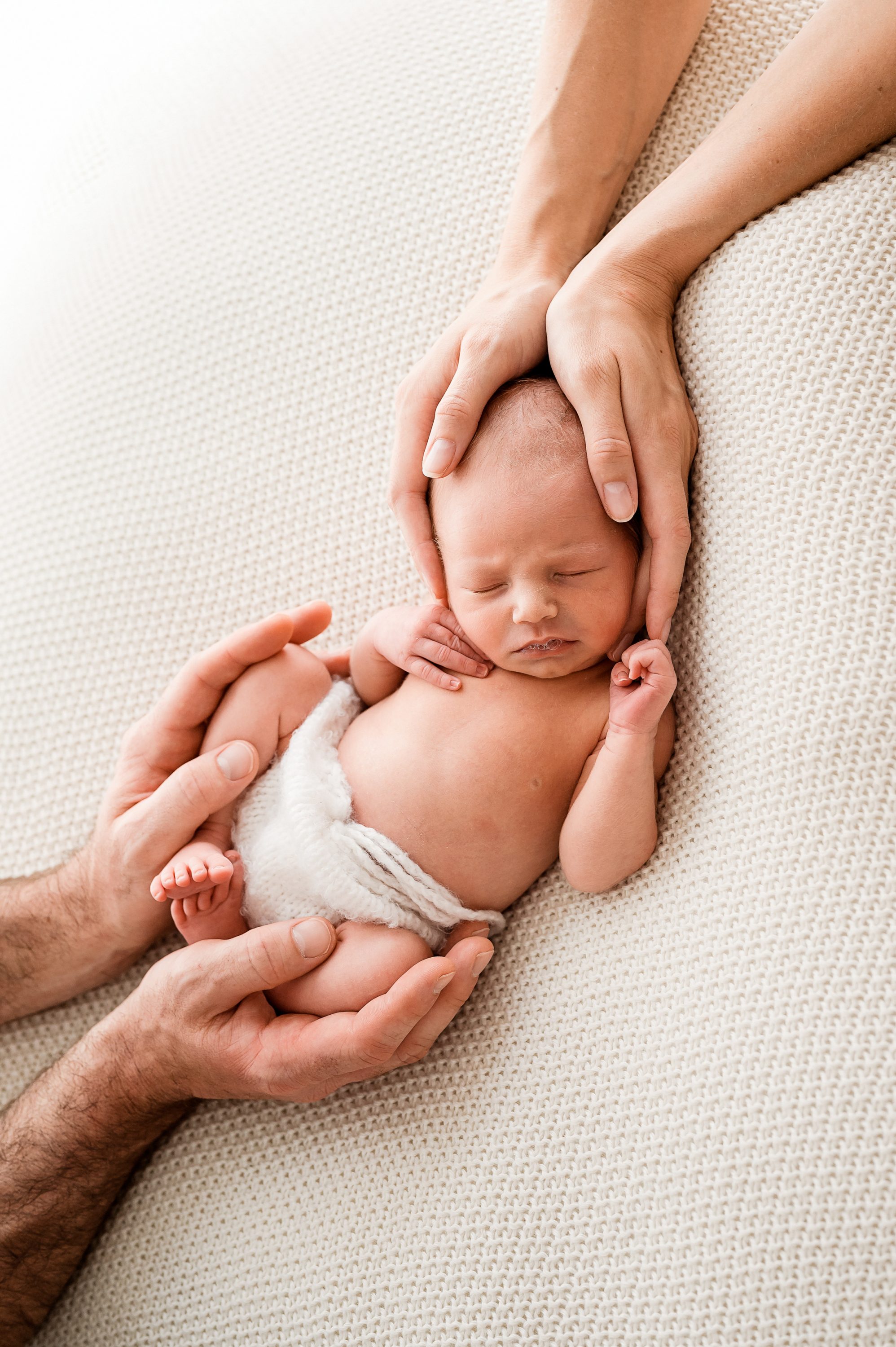 A newborn baby sleeps peacefully on a soft, cream-colored blanket while two adult hands gently cradle the baby's head and body—a timeless moment beautifully captured by a Family and Newborn Photographer Melbourne West