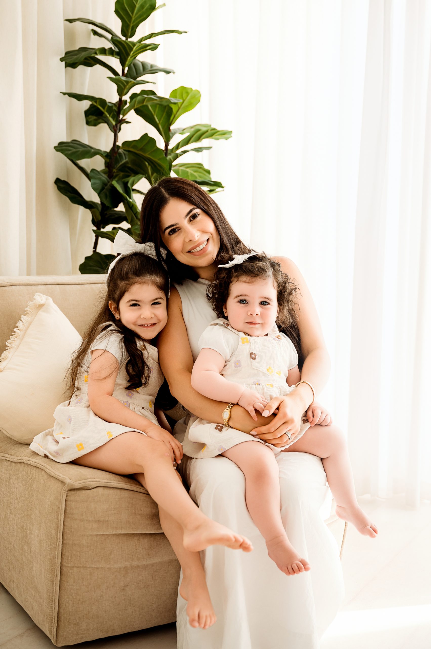 A woman sits on a beige sofa, smiling and holding two young girls—all dressed in light-colored outfits. A green plant and white curtains create a bright, cozy setting, perfect for a memorable Mother's Day mini session.