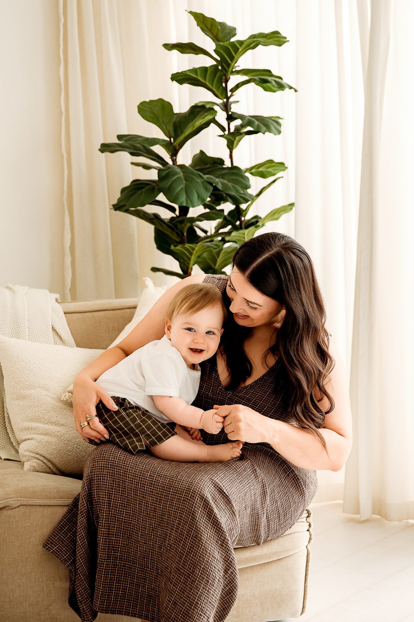 A woman with long dark hair sits on a beige sofa, smiling and holding a young child in her lap during a Mother’s Day mini session. The child, dressed in a white shirt and plaid shorts, is framed by sheer curtains and a large green plant in the background.