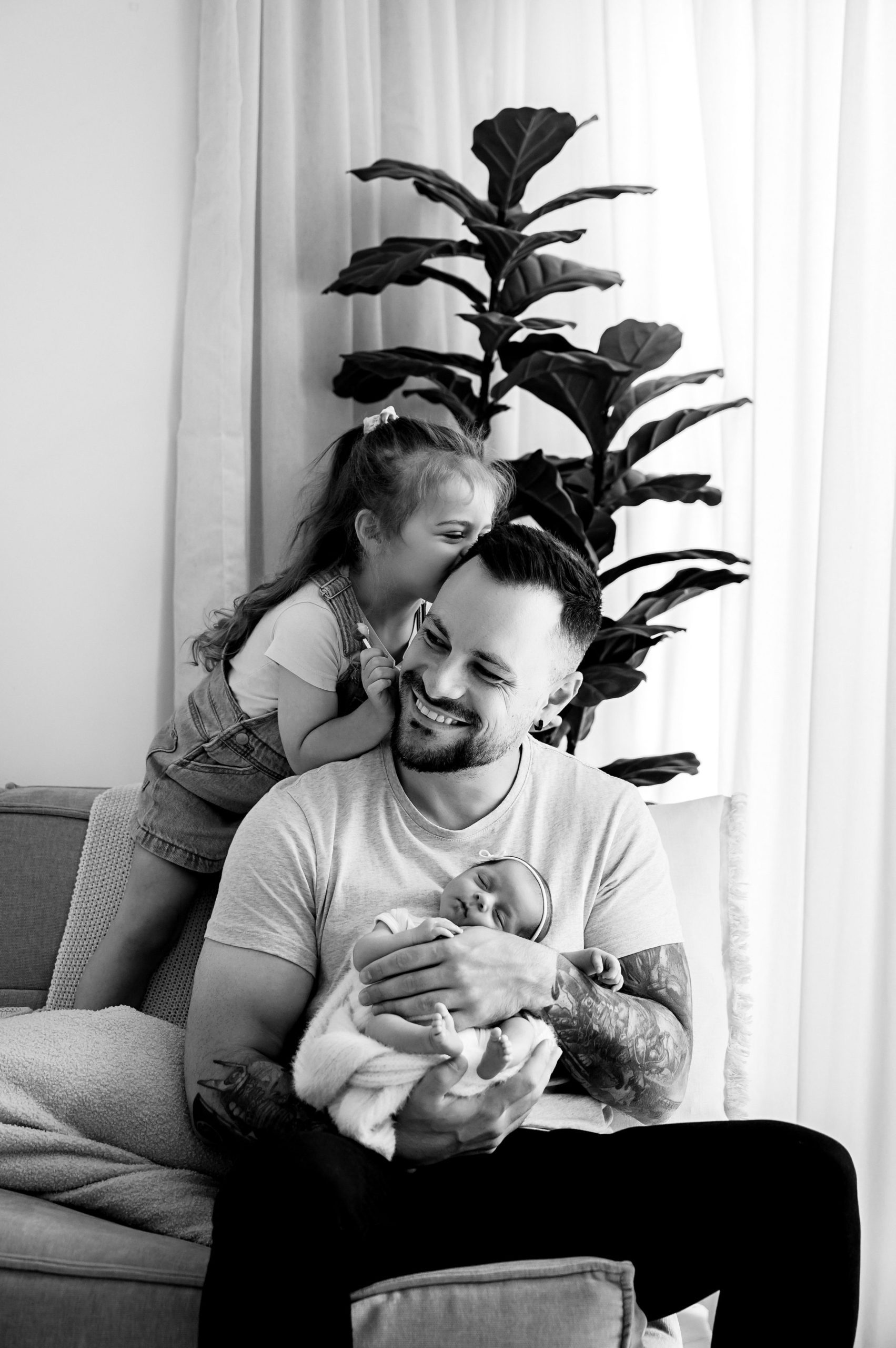 A man sits on a couch holding a baby and smiling, while a young girl whispers into his ear. Captured by a Family and Newborn Photographer Melbourne West , the scene features a large plant and curtains in the background.