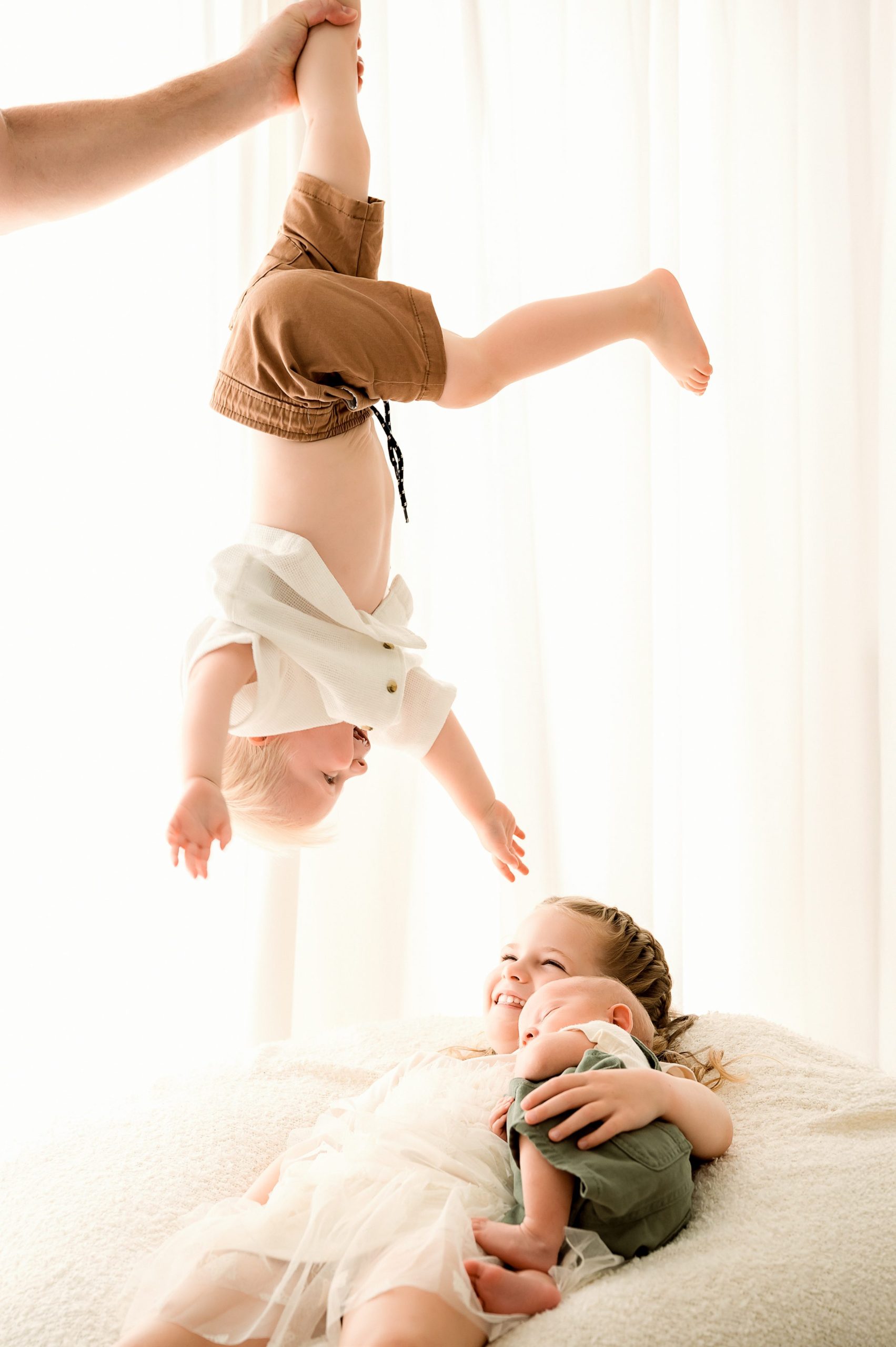 A toddler is held upside down by an adult hand, reaching joyfully toward a smiling young girl cuddling a newborn baby on a bed. Captured by a Family and Newborn Photographer Melbourne West , the bright scene glows with soft natural light.