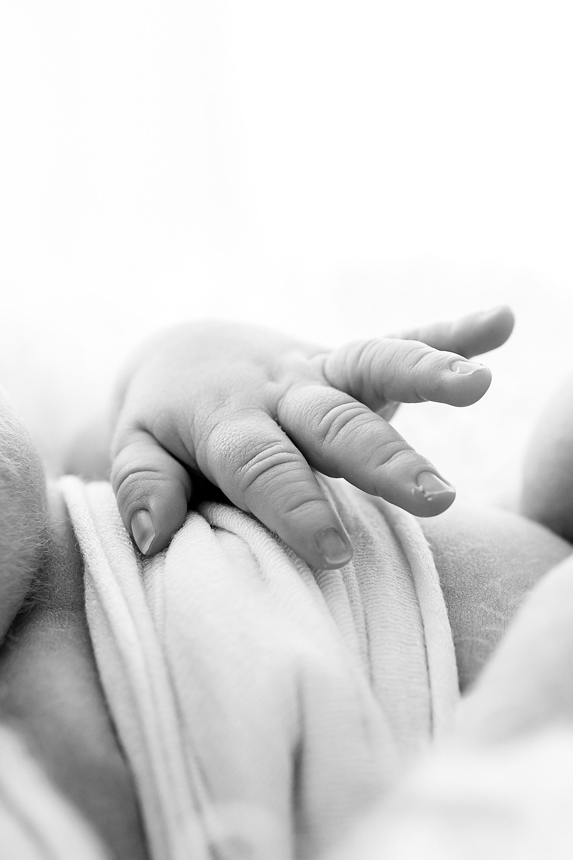 A close-up, black-and-white photo by a Family and Newborn Photographer Melbourne West captures a newborn baby's hand resting gently on a soft, white blanket, highlighting the tiny, delicate fingers and smooth skin.