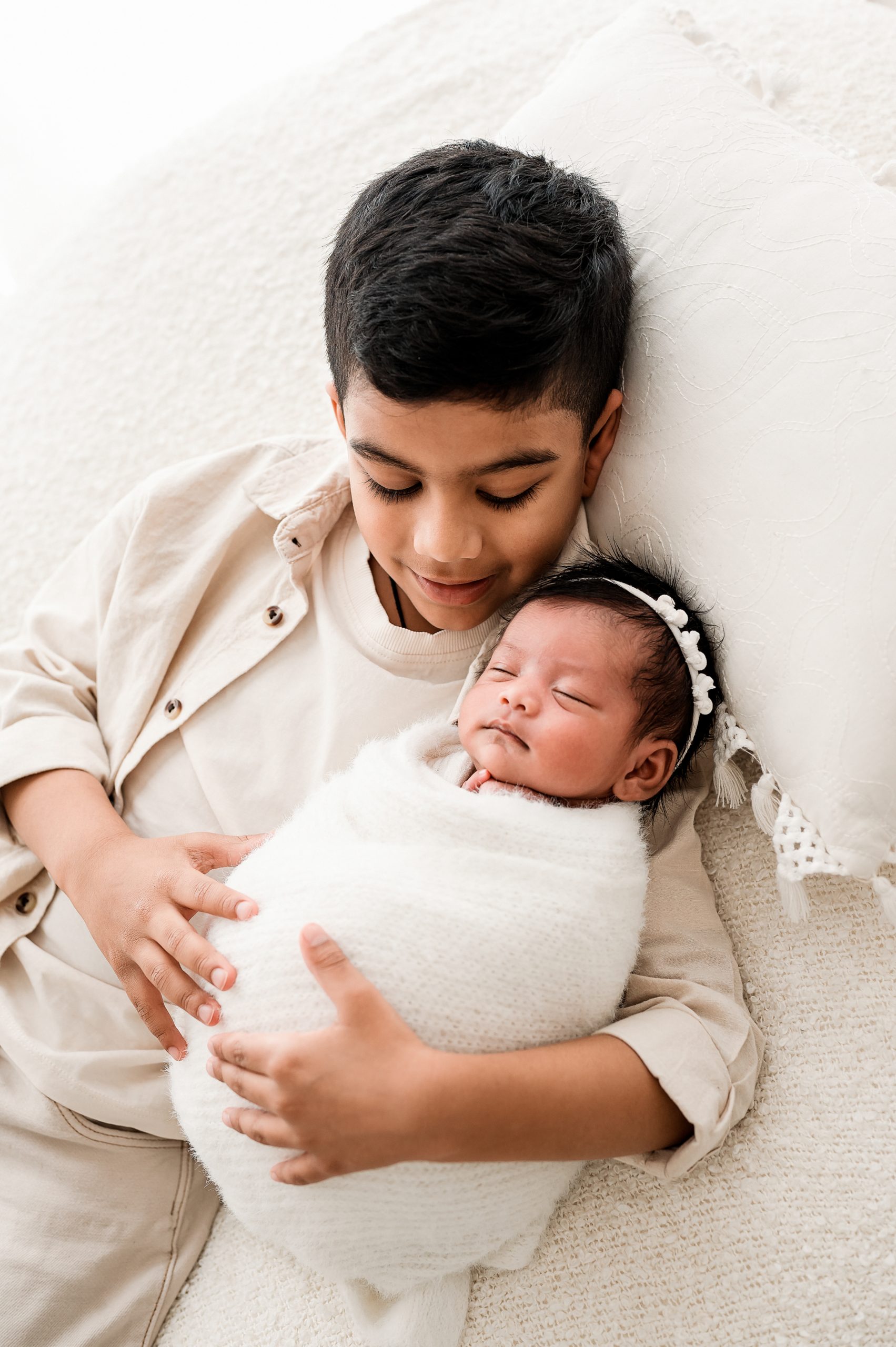A young boy smiles gently while cradling a sleeping newborn baby wrapped in a white blanket. Captured by a skilled Family and Newborn Photographer Melbourne West, both rest on a soft, white surface, cherishing this precious moment.