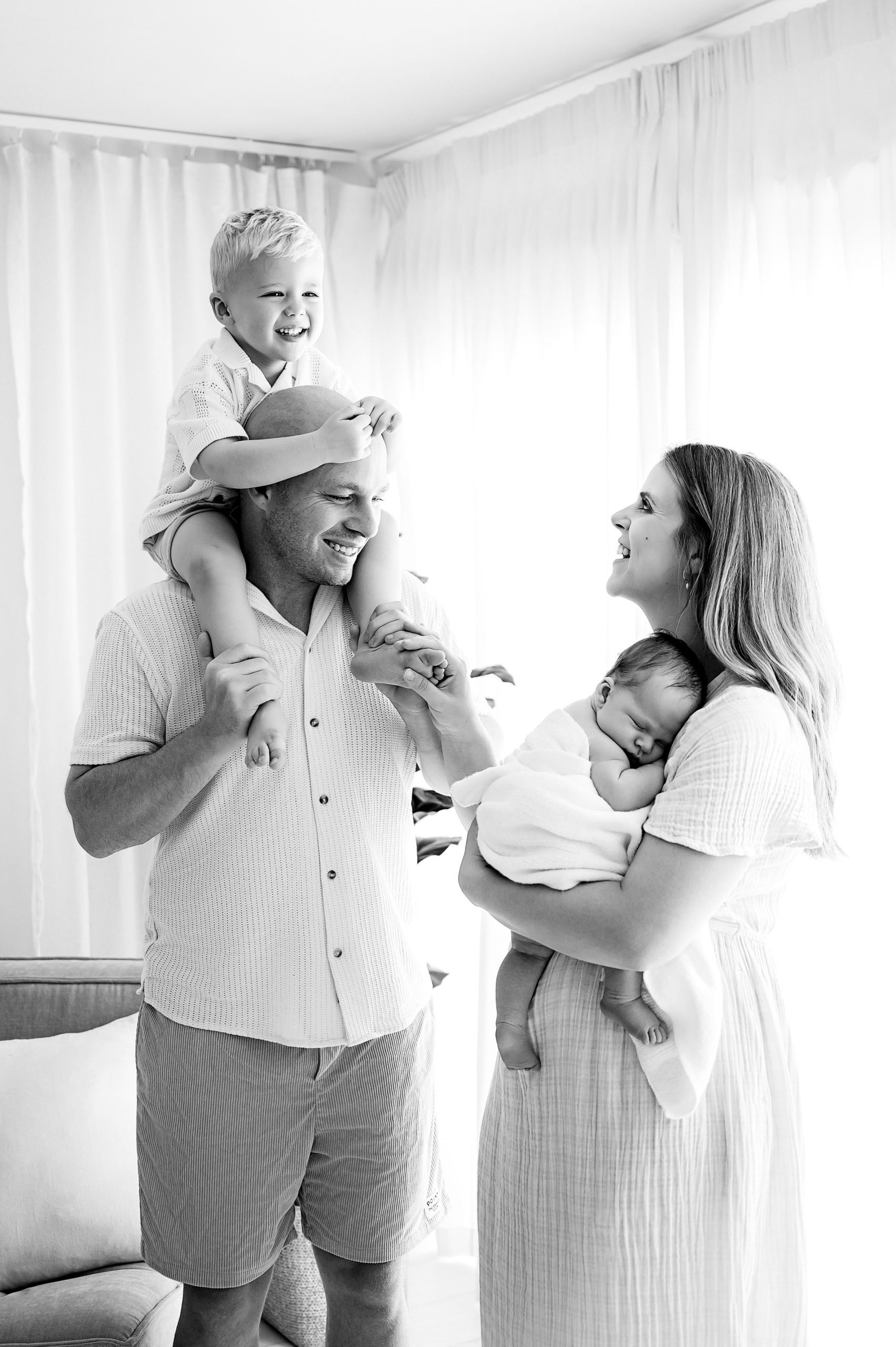 A man smiles with a young boy on his shoulders while a woman holding a baby grins at them, capturing what to expect from joyful family moments by a Family and Newborn Photographer Melbourne West. The family stands in a bright room with sheer curtains and soft natural light.