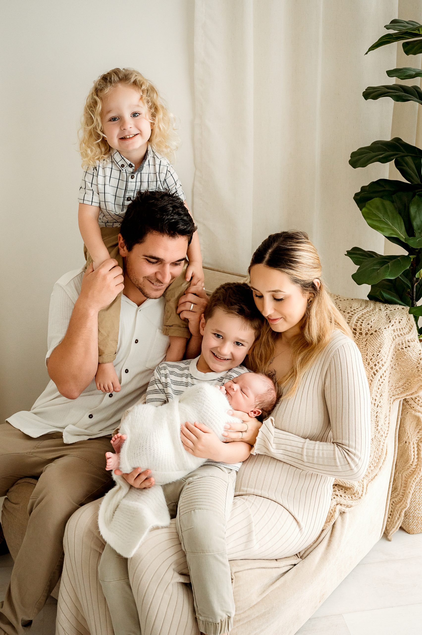 A family of five poses indoors: a woman in a cream dress holds a swaddled newborn, with the help of a talented Family and Newborn Photographer Melbourne West, as they all smile warmly together.