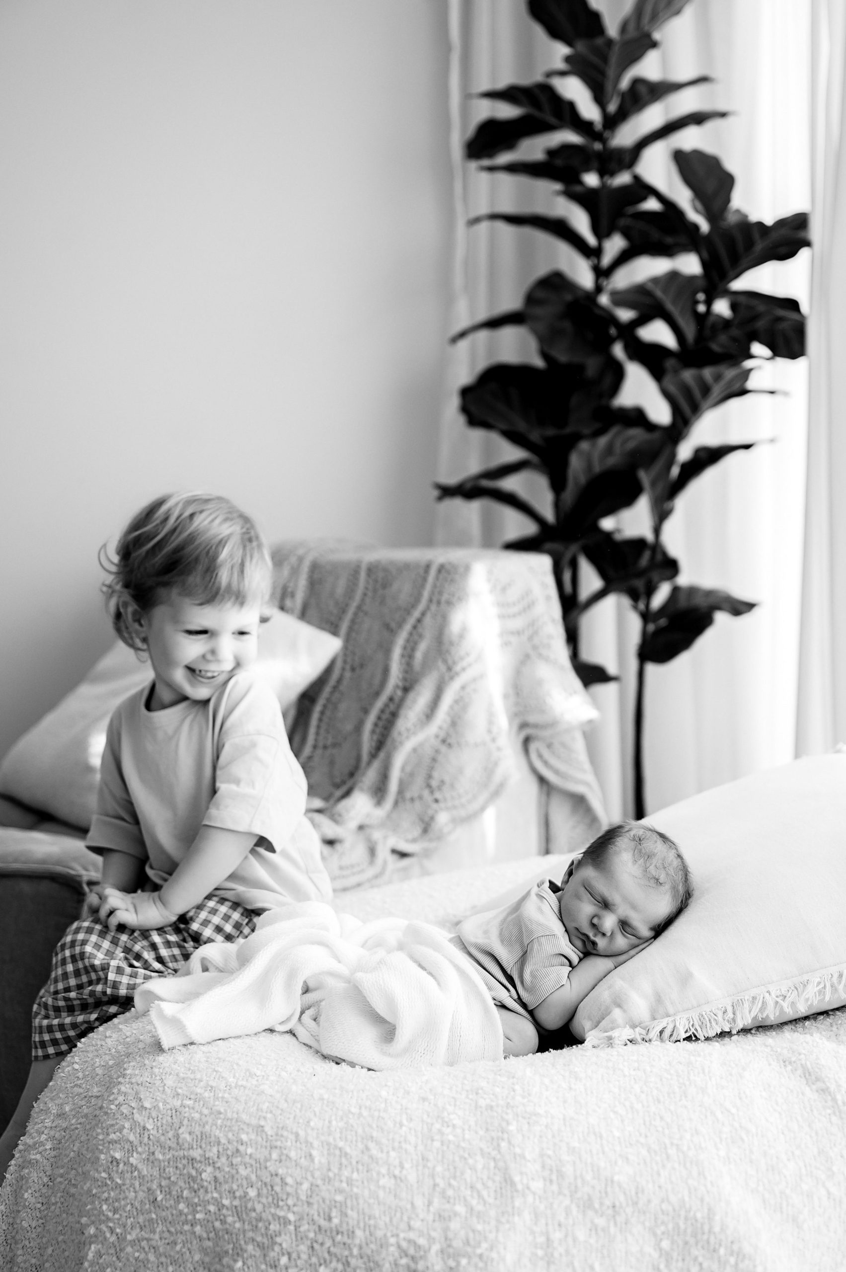 A young child smiles while sitting on a bed next to a sleeping newborn wrapped in a blanket, with a cozy throw and tall plant in the background—a timeless black and white moment by a Family and Newborn Photographer Melbourne West