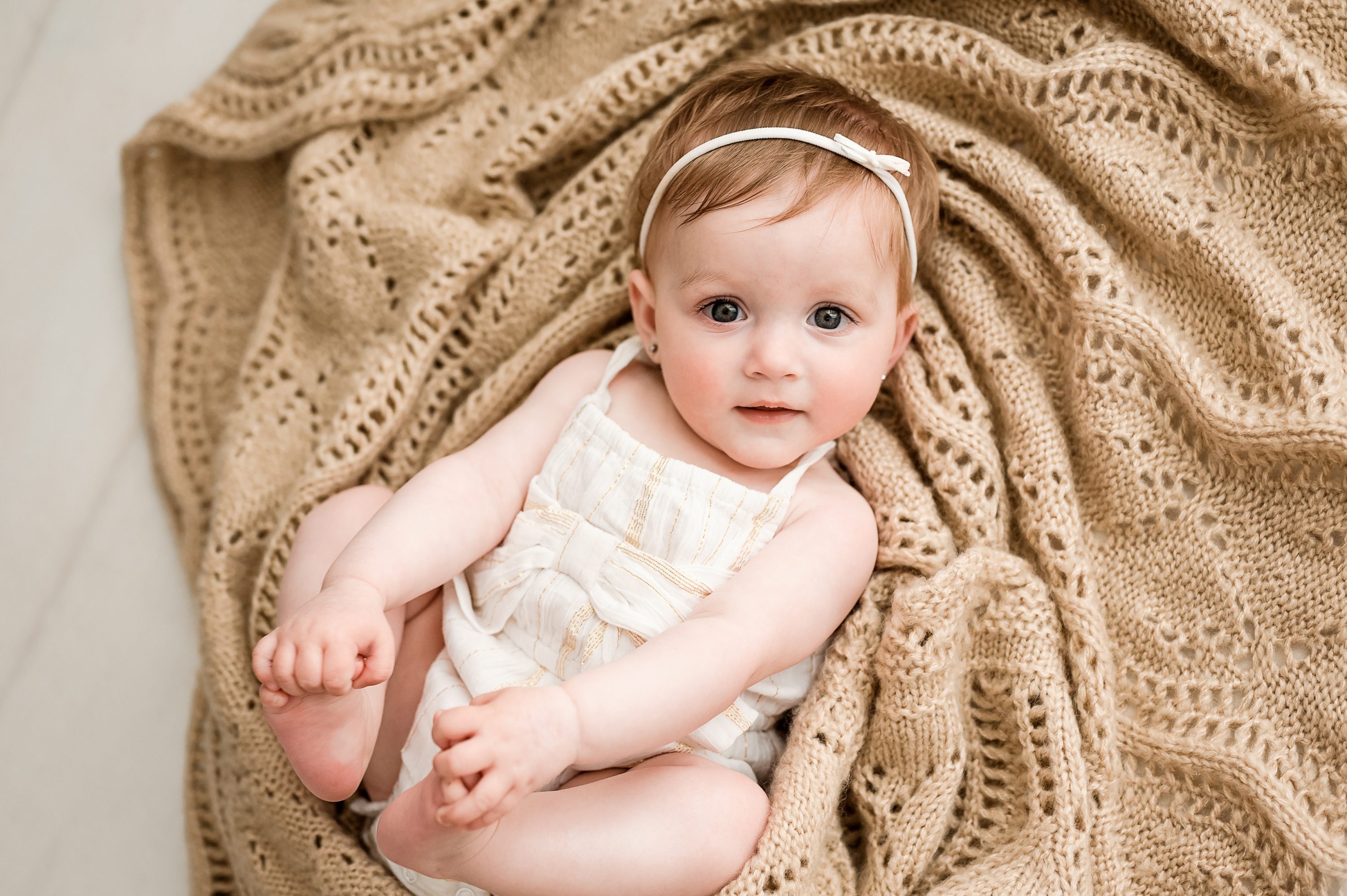 A baby with light brown hair and a white headband sits on a textured beige blanket, looking up at the camera with big eyes, wearing a sleeveless light-colored outfit—captured by a Family and Newborn Photographer Melbourne West