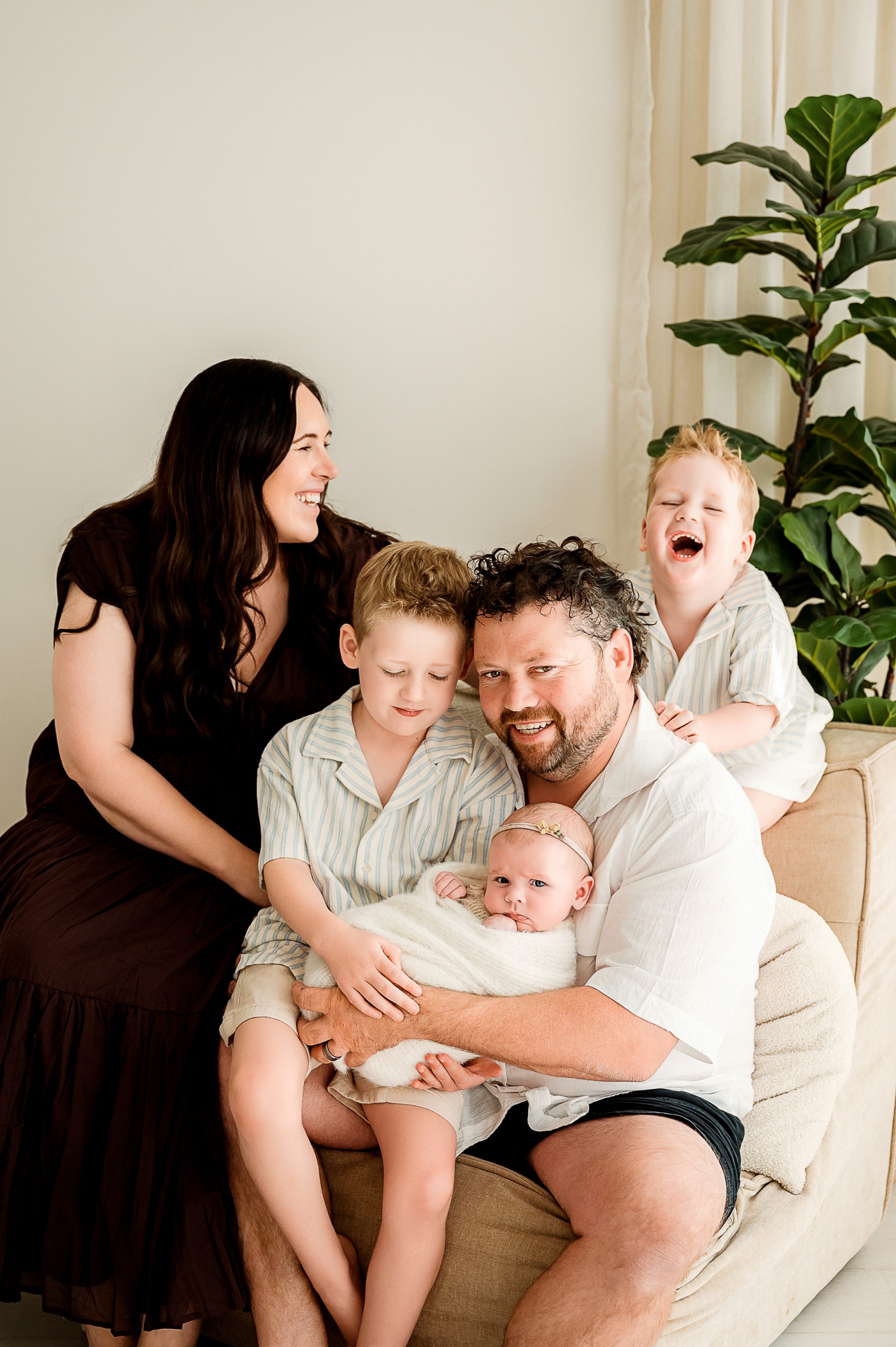 A family of five poses together indoors on a beige couch. The mother and father smile, with the father holding a baby. Captured by a Family and Newborn Photographer Melbourne West , two young boys are beside them, one laughing joyfully. A green plant is in the background.