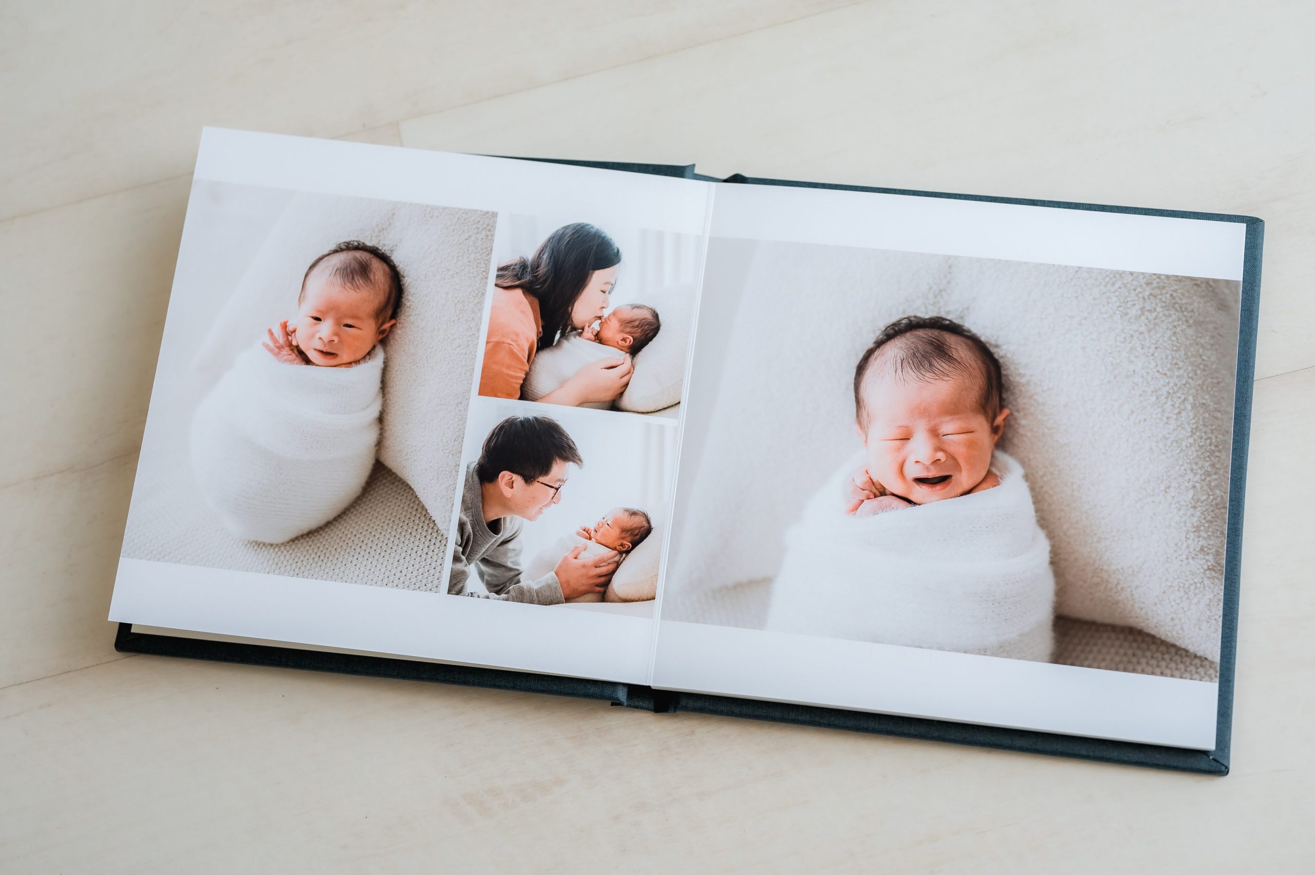 An open heirloom album displays newborn photography Melbourne style, featuring a baby wrapped in a white blanket and two adults interacting lovingly in some of the photos.