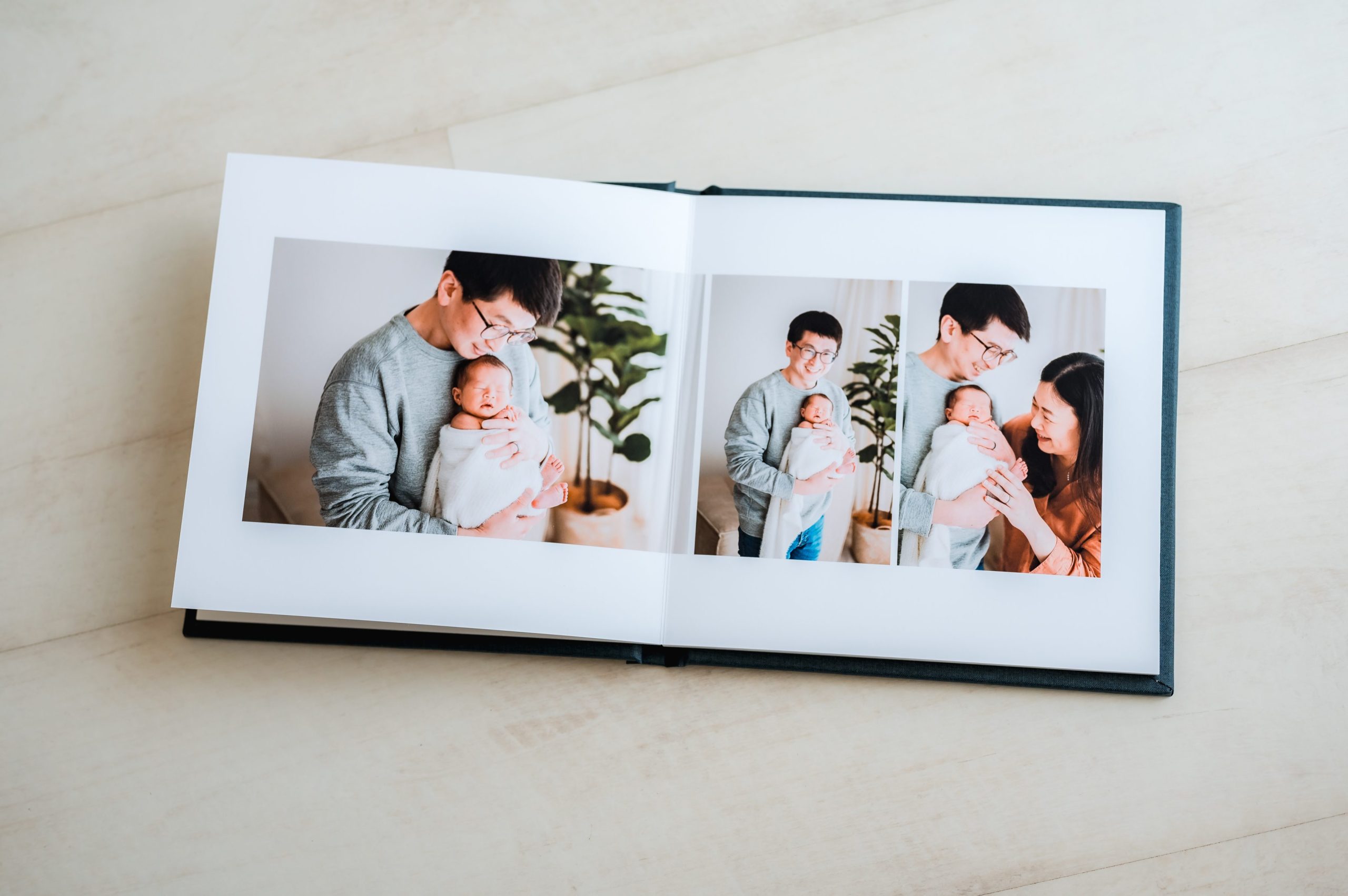 An open heirloom album on a light surface displays three photos of two adults holding and smiling at a newborn baby, with a potted plant visible in the background.