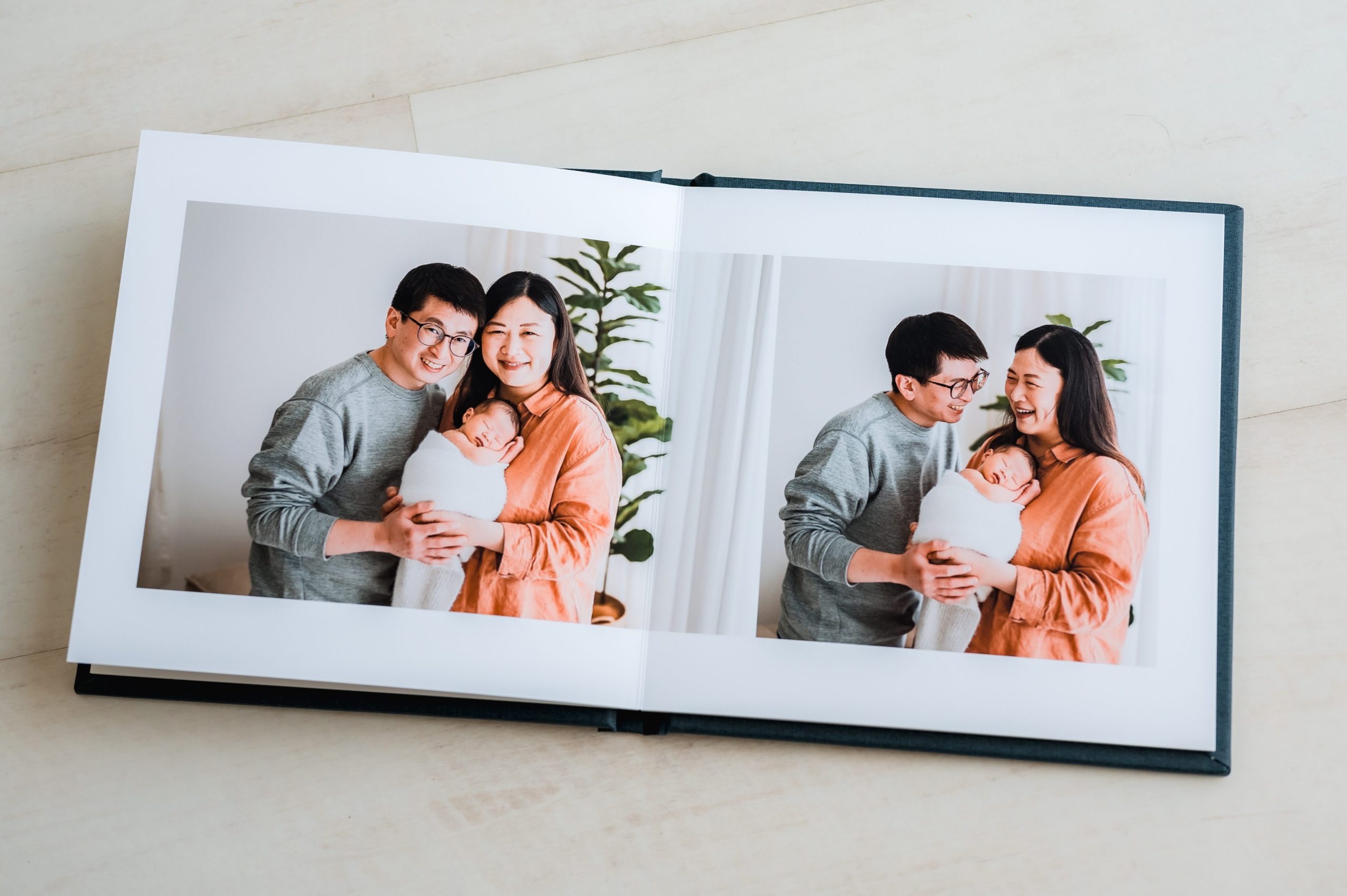 An open heirloom album displays two pictures of a smiling couple holding a baby wrapped in a white blanket, standing together indoors with a green plant in the background—capturing timeless moments of newborn photography Melbourne.