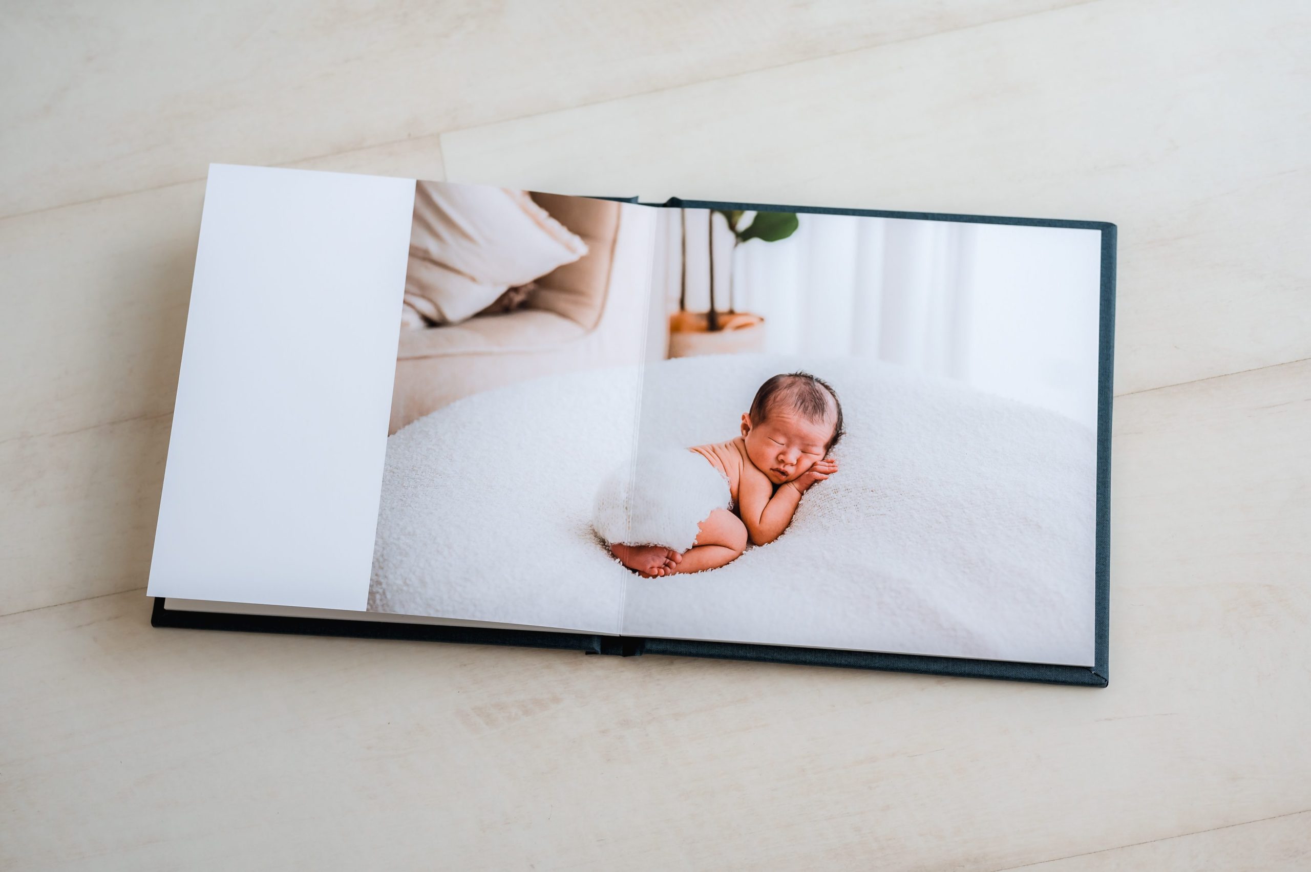 An open heirloom album lies on a light-colored surface, displaying a photo of a newborn baby sleeping on a white blanket in a cozy room with soft, natural lighting—perfect for those seeking timeless newborn photography Melbourne memories.