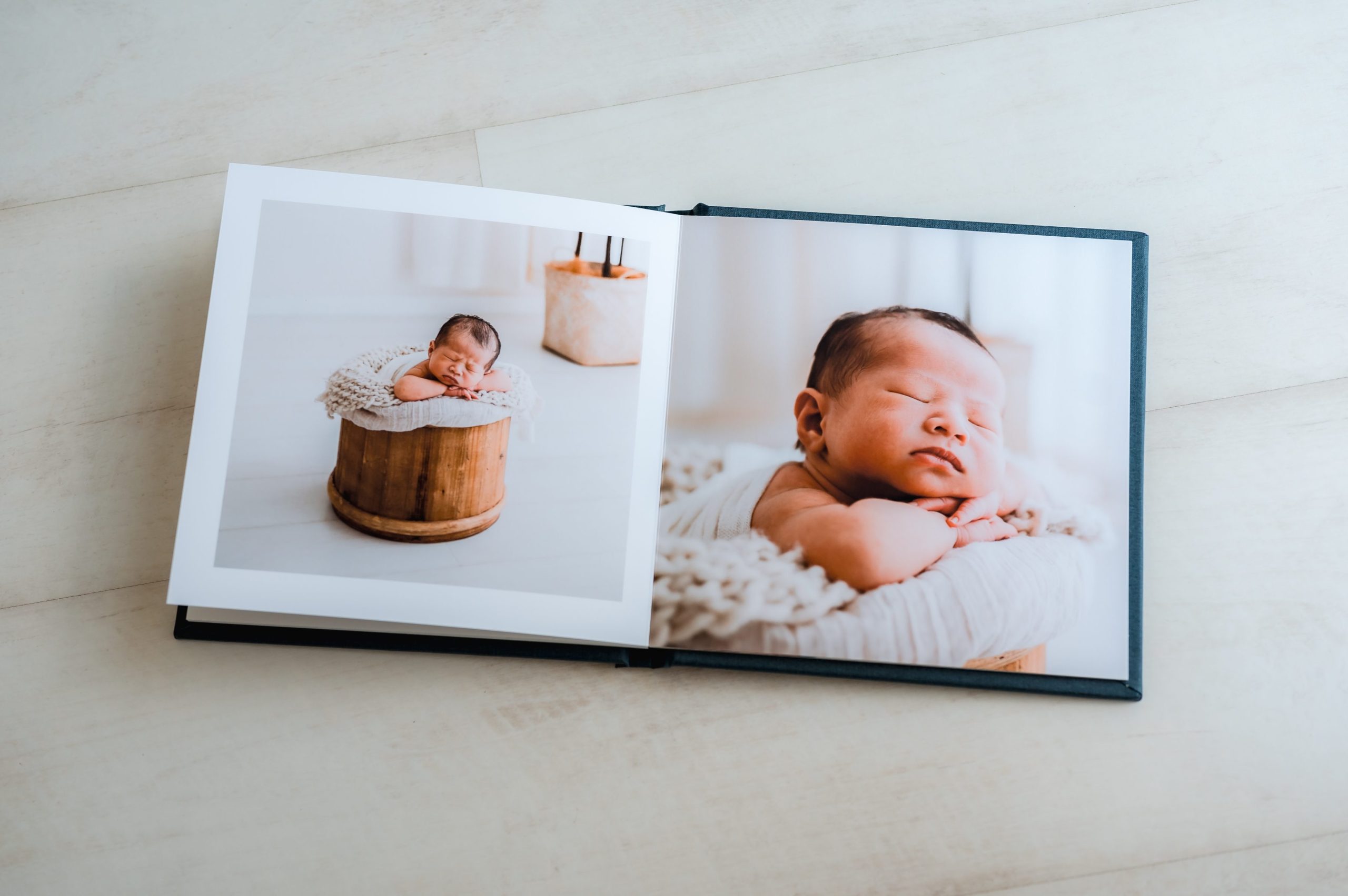 An open heirloom album showcases newborn photography Melbourne-style with two photos: a baby sleeping in a wooden bucket on a blanket, and a close-up of the newborn resting on a soft surface.
