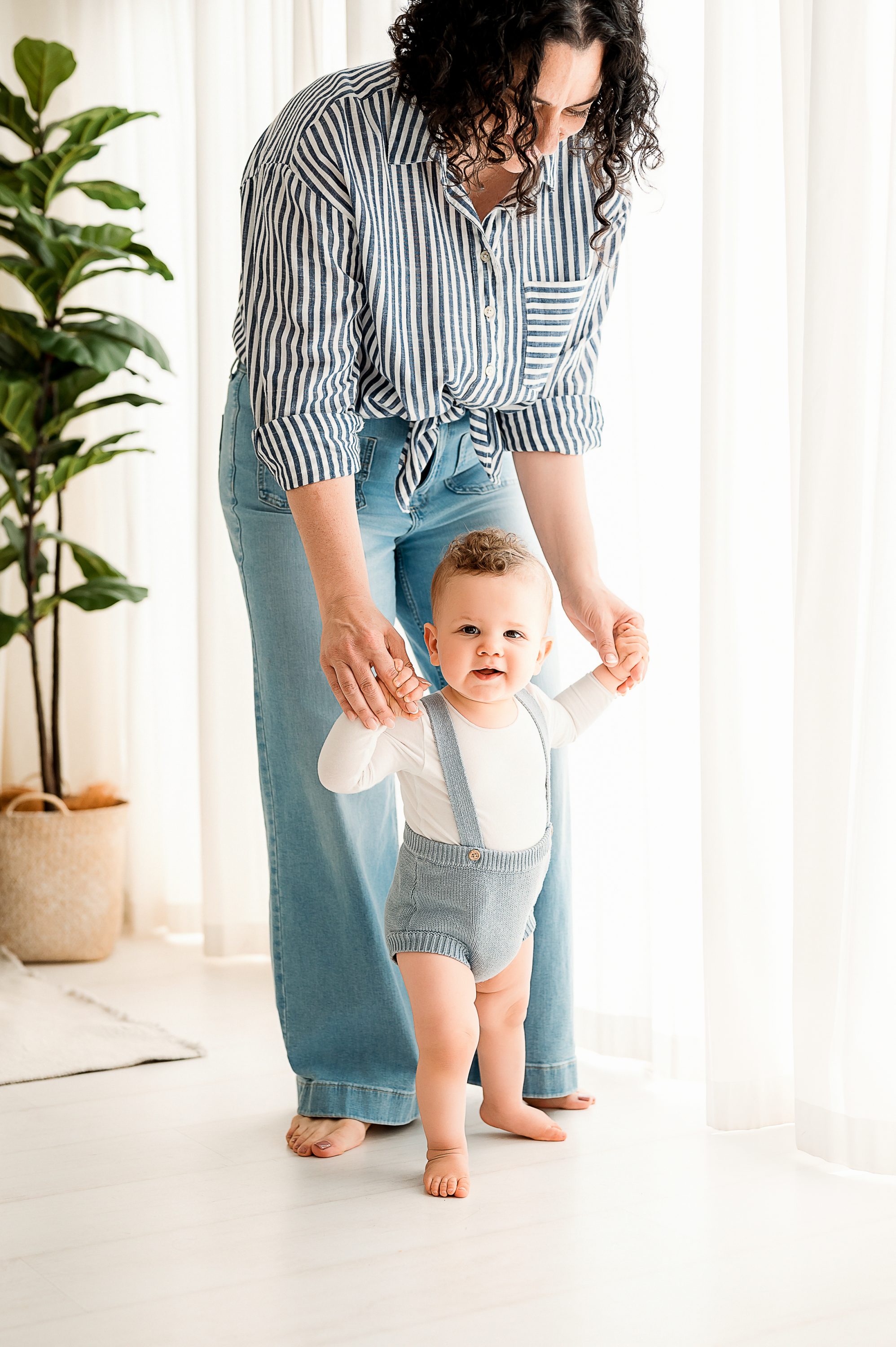 An adult helps a toddler stand and walk by holding their hands. Captured by a Family and Newborn Photographer Melbourne West, the scene features a bright room, a plant by the window, and warm moments in stylish outfits.