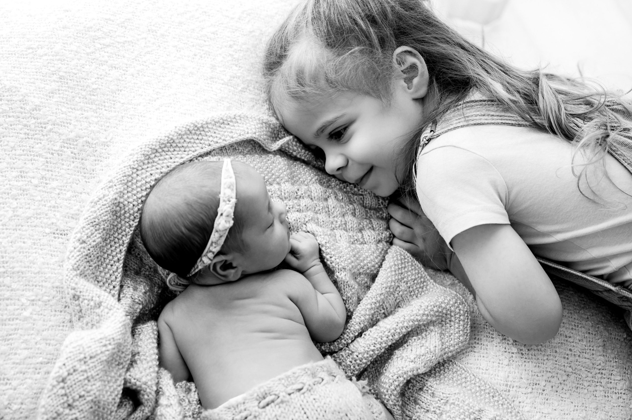 A young girl with long hair smiles lovingly at a newborn baby wrapped in a blanket. Perfect for newborn photography Melbourne, the baby wears a headband as both children lie on a soft textured surface in this black and white image.