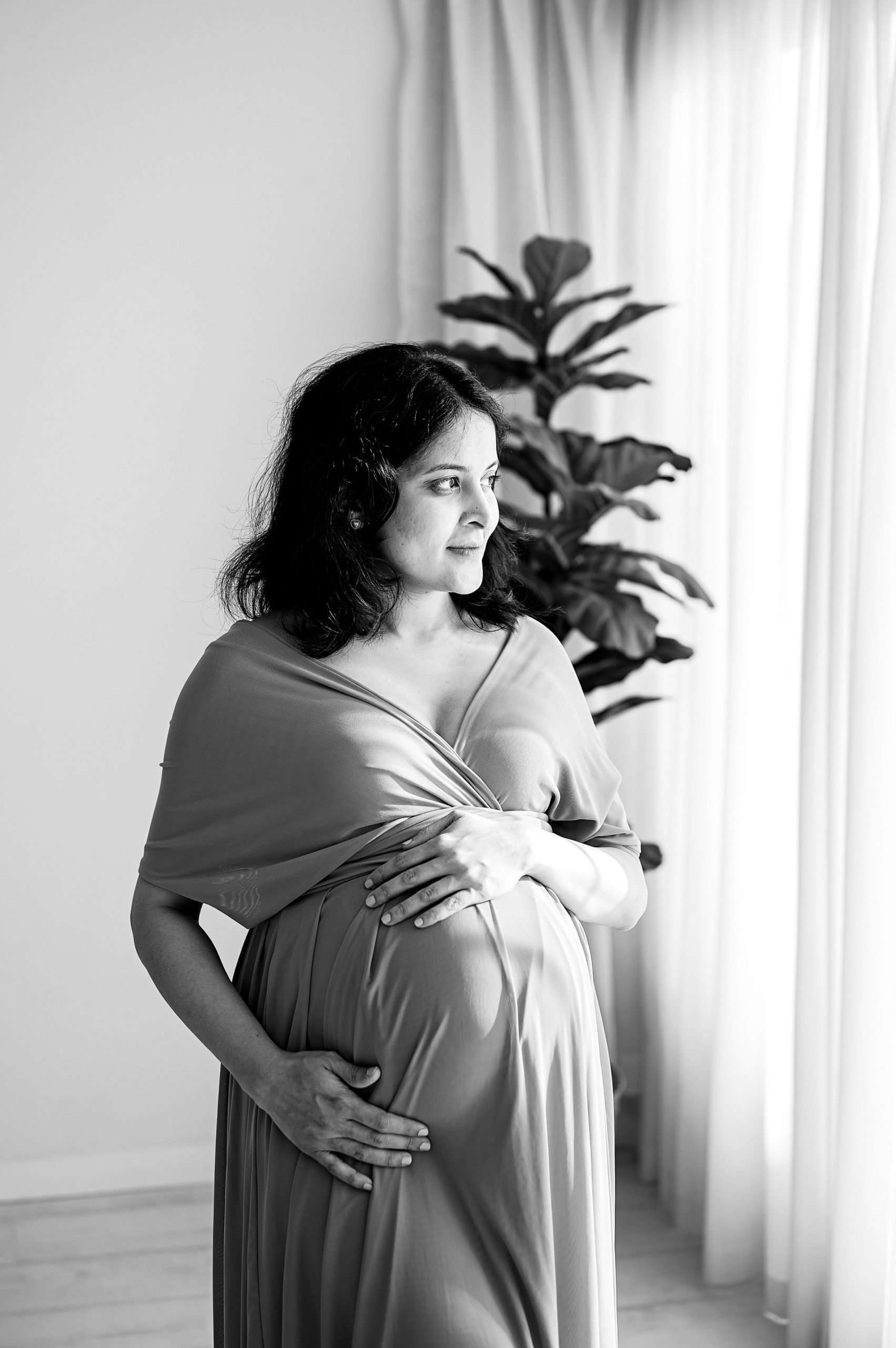 A pregnant woman in a flowing dress stands indoors by a window, gently cradling her belly with both hands and looking thoughtfully into the distance. A leafy plant is visible in the background. Black and white maternity photography Melbourne West.