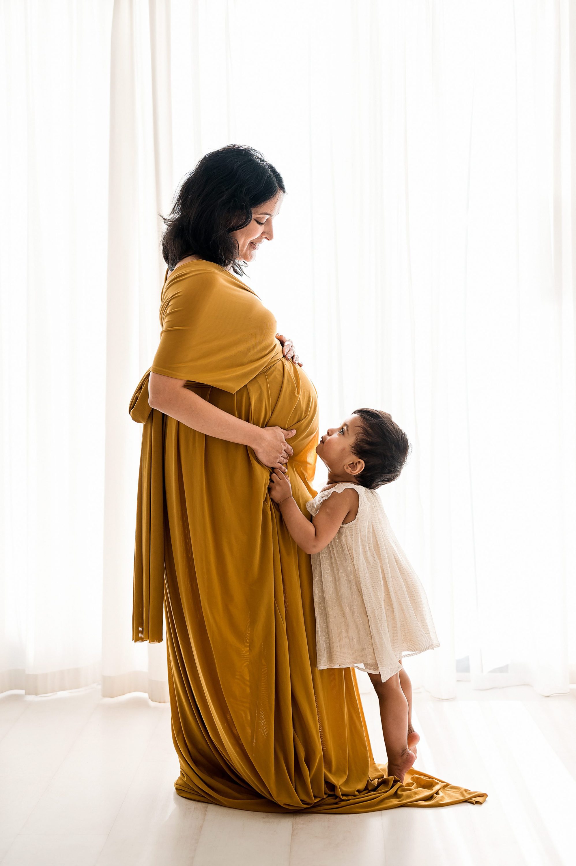 A pregnant woman in a mustard yellow dress stands by a bright window, smiling down at a young child in a white dress who is touching her belly—capturing the warmth of Maternity Photography Melbourne Westt.