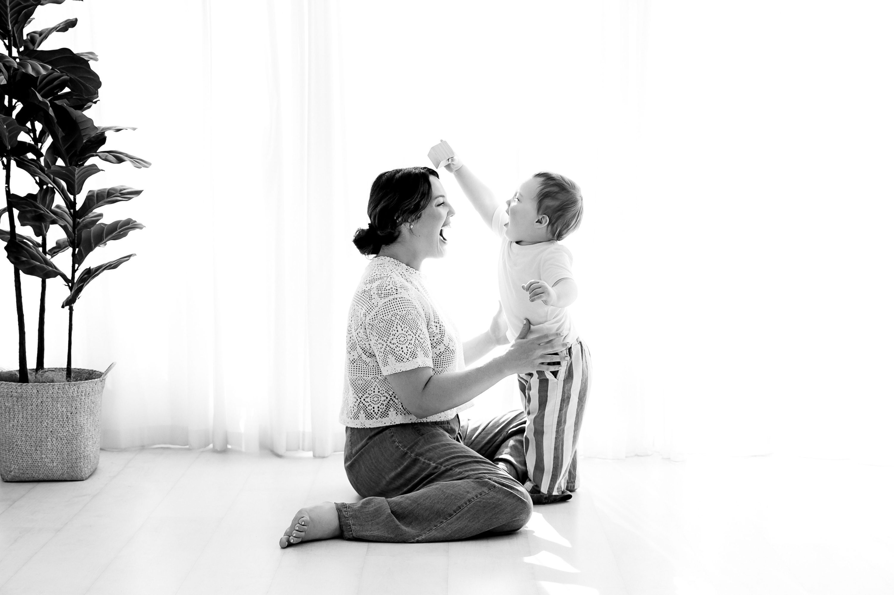 A woman sits on the floor, smiling at a standing toddler who raises one arm excitedly. In a bright room with sheer curtains and a potted plant, this tender moment is captured by a Family and Newborn Photographer Melbourne West