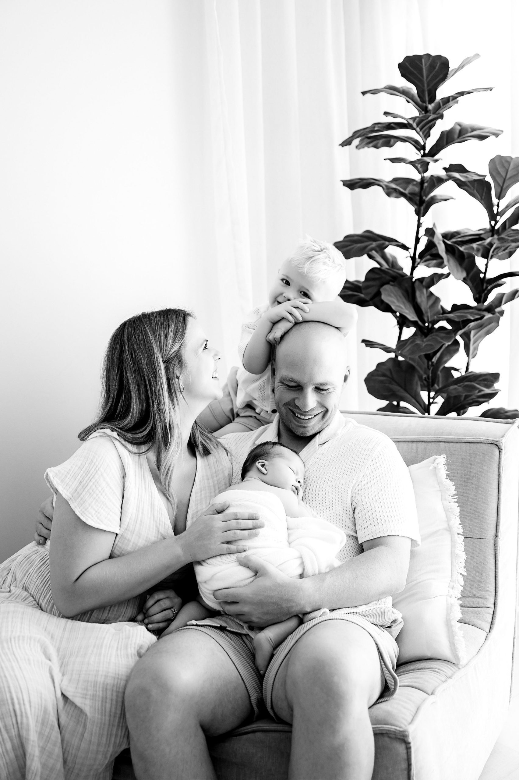 A smiling family of four sits on a couch in this black and white family and newborn photography scene. The mother and father hold a newborn, while a toddler sits on the father's shoulders, touching his head. A large plant is in the background.