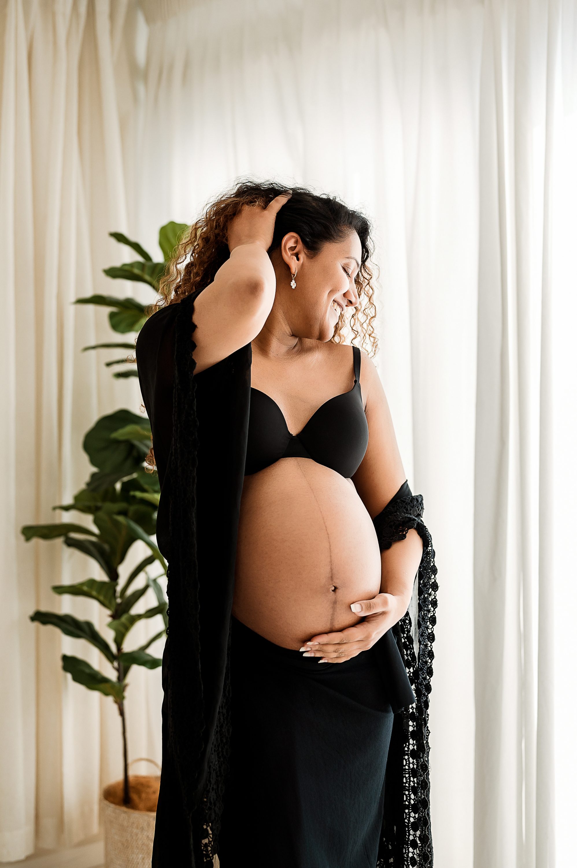 A smiling pregnant woman in a black bra and draped fabric stands indoors by a window with sheer curtains, holding her belly and touching her hair. A green potted plant is in the background—perfect for Maternity Photography Melbourne West.