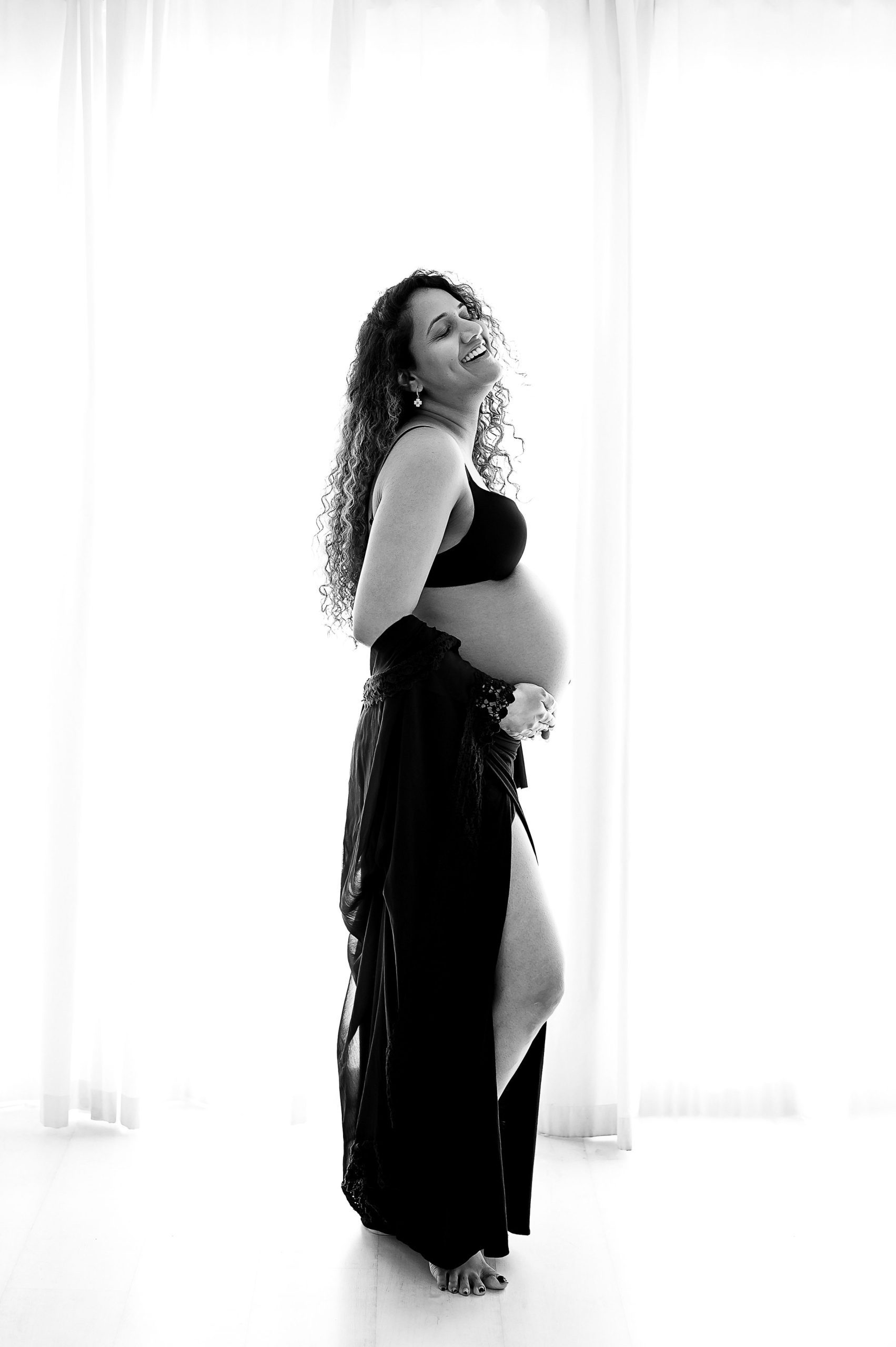 A pregnant woman with curly hair smiles and poses in profile, wearing a black bra and sheer skirt, standing barefoot in a bright, softly lit room with white curtains—capturing the essence of maternity photography Melbourne West.