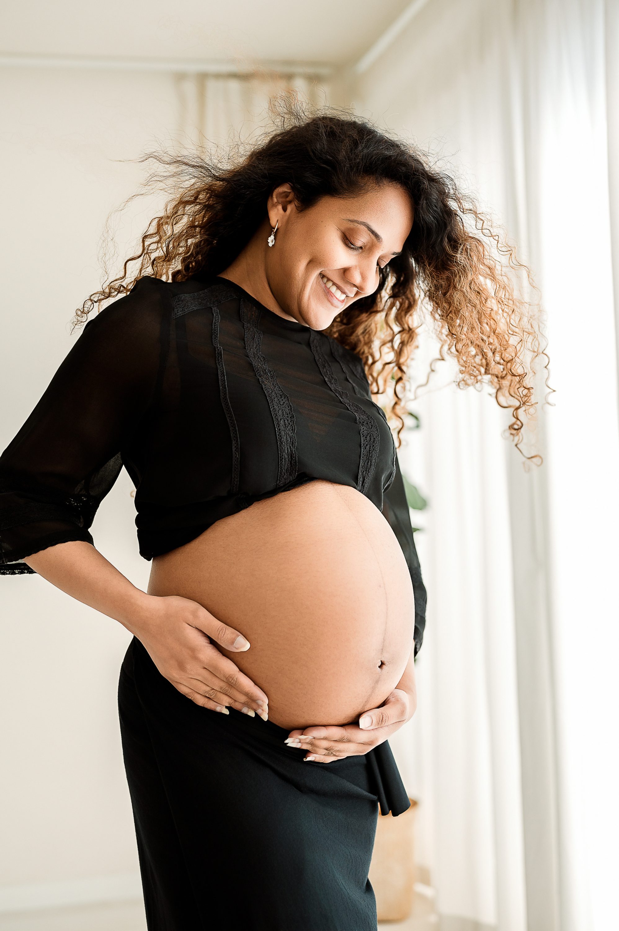 A pregnant woman with curly hair smiles and looks down while cradling her bare baby bump with both hands. Perfect for maternity photography Melbourne West, she wears a sheer black top and skirt in a bright, softly lit room.