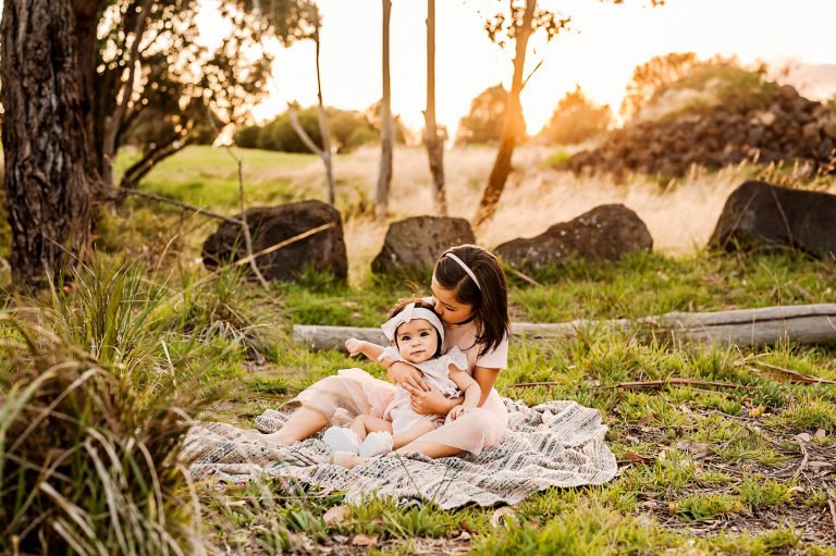 Two young children sit on a blanket in a grassy outdoor setting at sunset—one hugging the other, surrounded by trees and rocks. It's a perfect example of the charm found at family photo locations in Melbourne's West, bathed in warm sunlight.