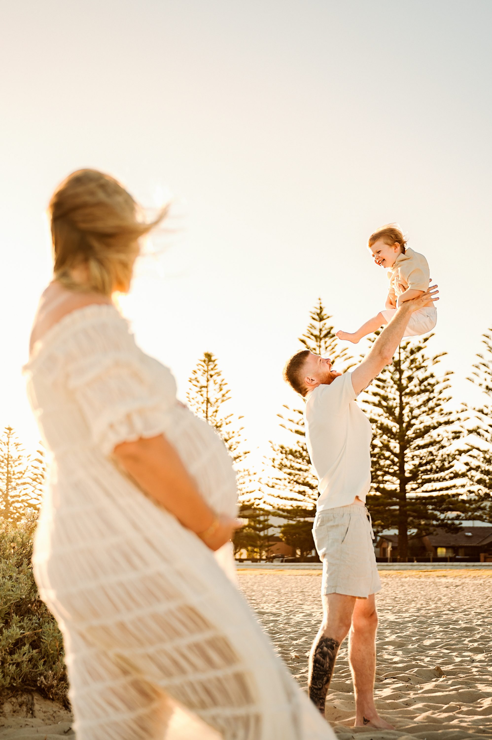 A pregnant woman in a white dress stands on a sandy beach, watching as a man lifts a smiling child into the air. Perfect for maternity photography Melbourne West, the scene glows with tall trees and a warm sunset in the background.