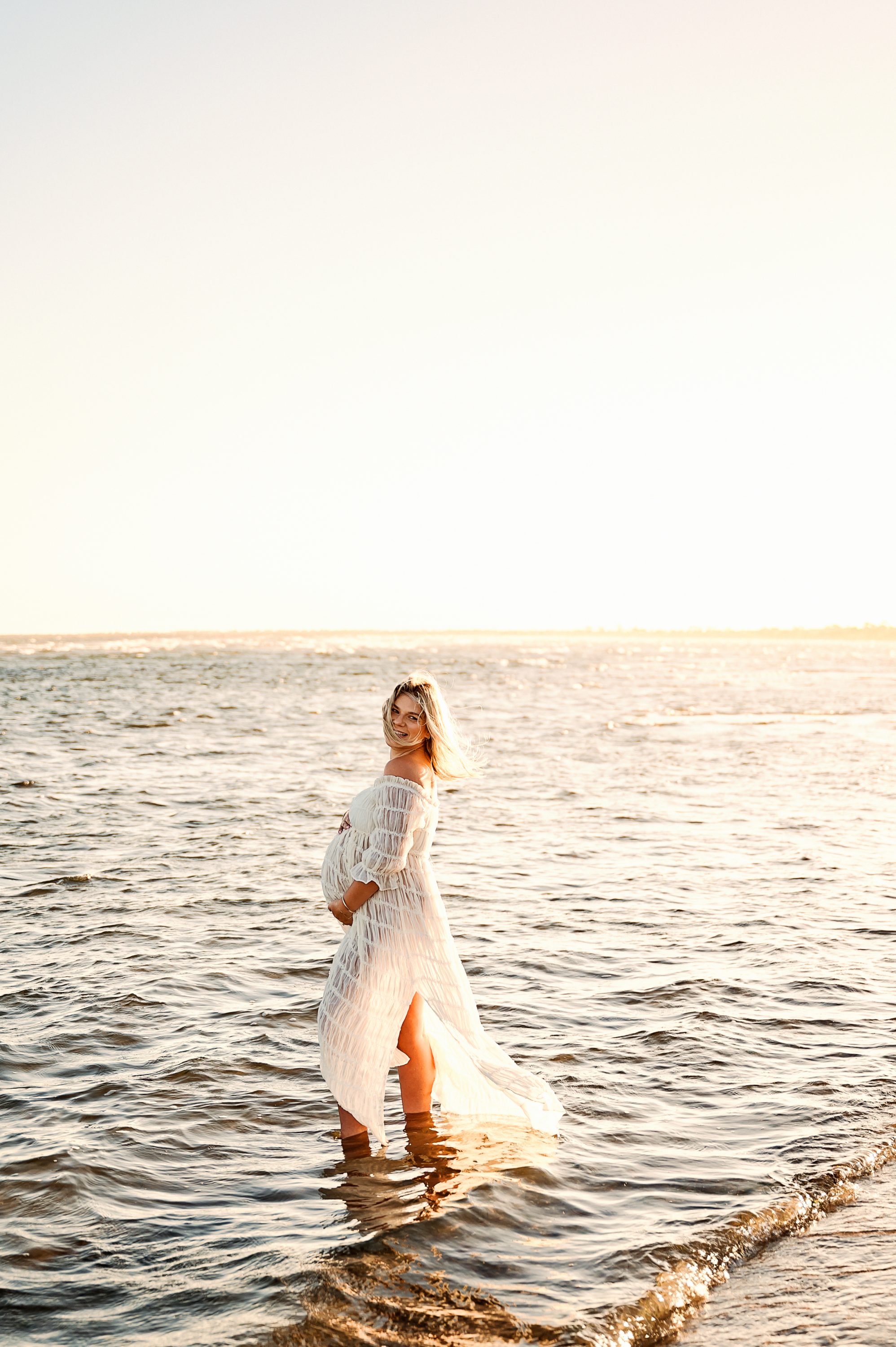 A woman in a long, white, off-shoulder dress stands in shallow ocean water at sunset, smiling and holding her baby bump. Gentle waves surround her feet as the sky glows warmly—perfect for maternity photography Melbourne west.