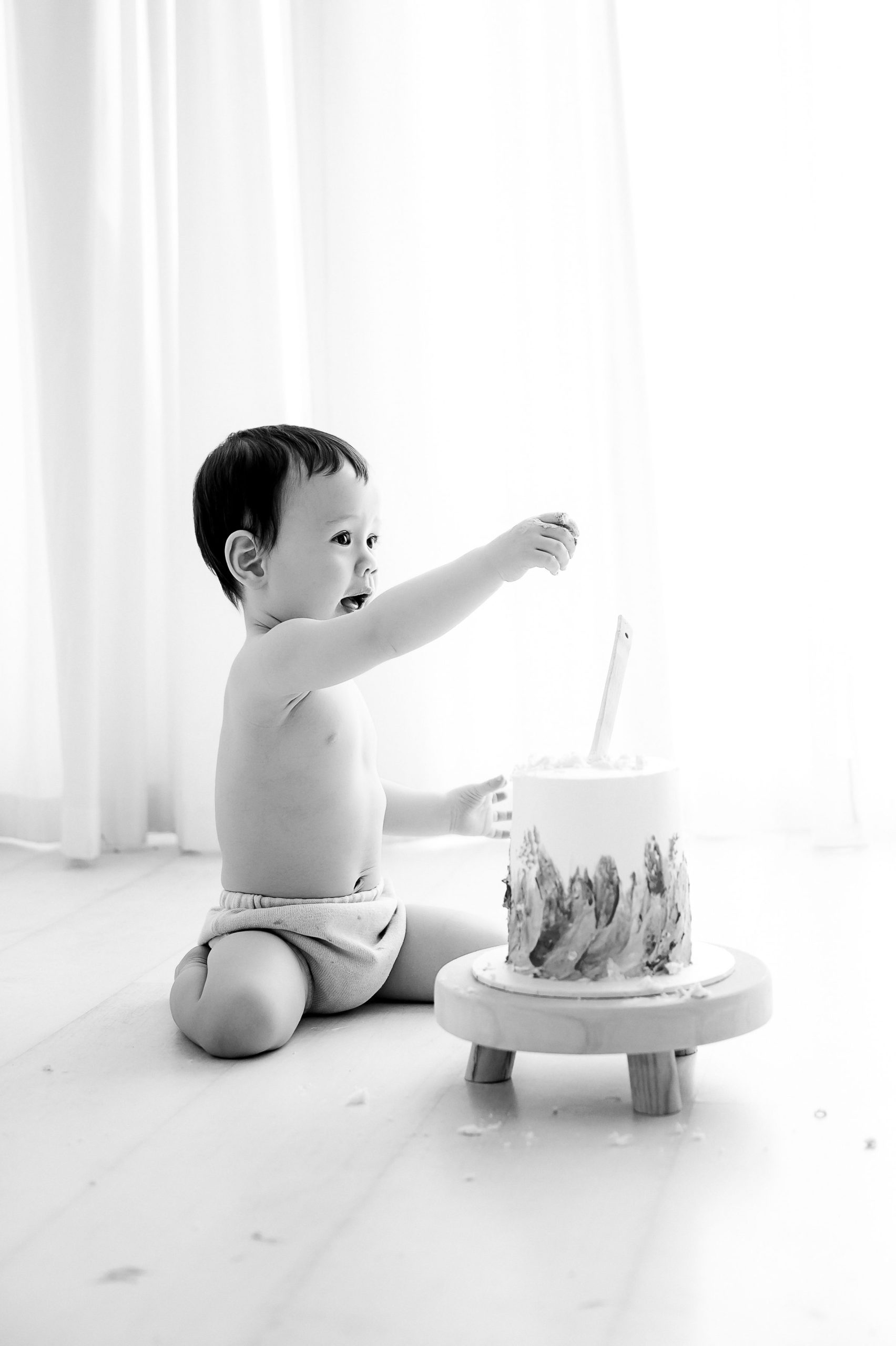 A baby sits on the floor in front of a cake, reaching out with excitement. Perfect for cake smash photography Melbourne, the shirtless little one in a diaper is surrounded by bright, airy light and soft curtains in the background.
