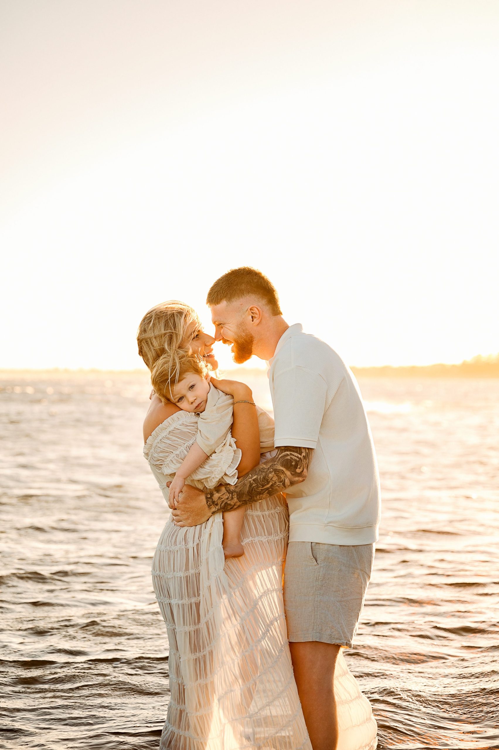 A man and woman stand in shallow water at sunset, embracing and holding a small child on Mum's pregnant belly. The scene is warmly lit by the golden sun—an intimate moment beautifully captured in a maternity photography Melbourne West session.