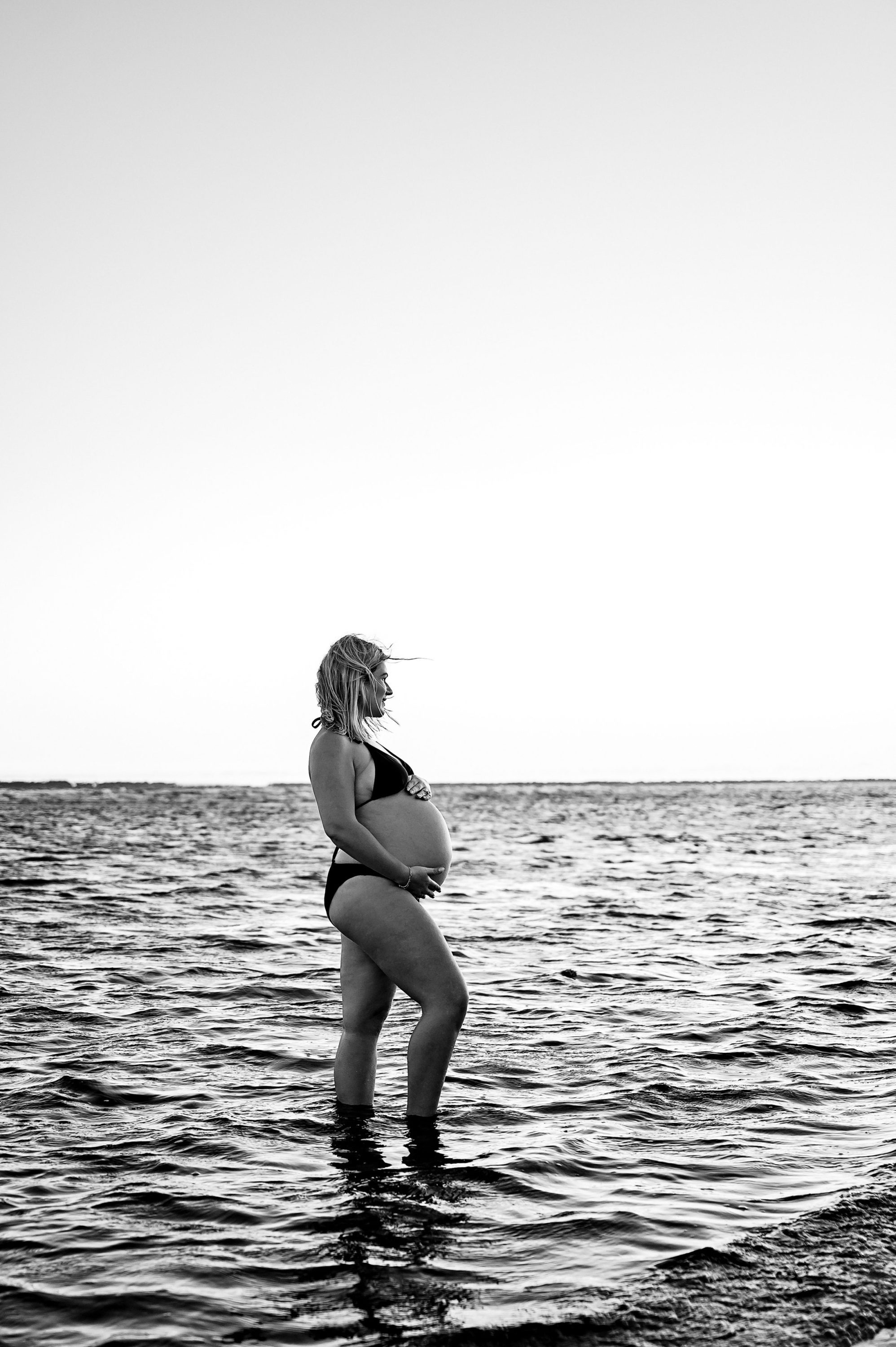 A pregnant woman in a bikini stands in shallow ocean water, gazing to the side. This black and white scene, with a calm sea and clear sky, beautifully captures the essence of maternity photography Melbourne west.