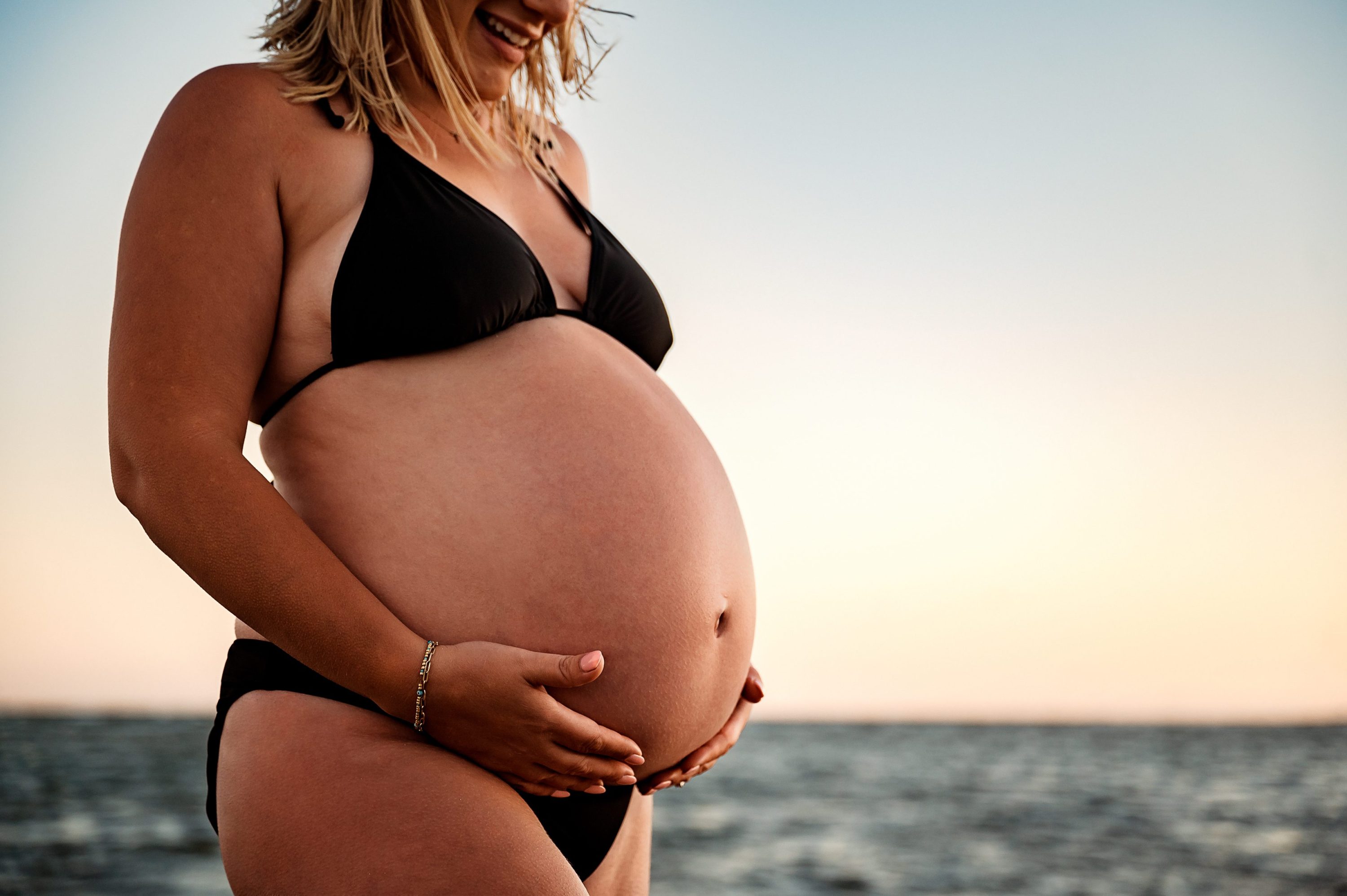 A pregnant woman in a black bikini stands on a beach, smiling and gently holding her belly, with the ocean and a sunset sky in the background—perfect for maternity photography Melbourne West.