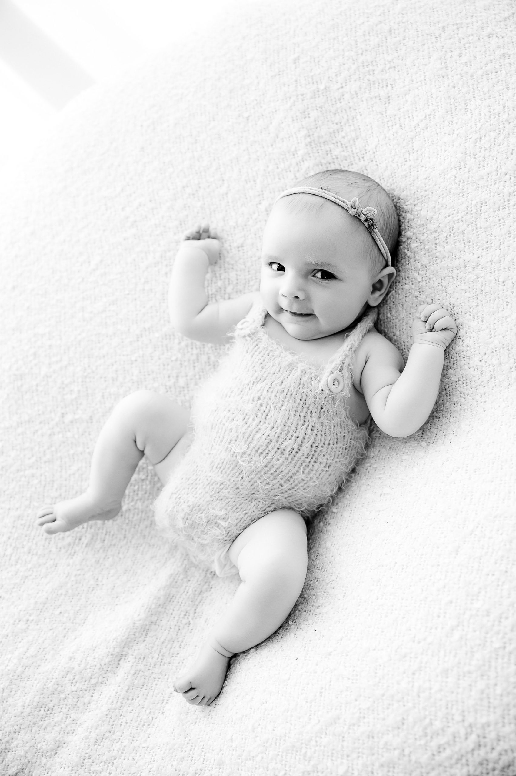 A baby wearing a knitted outfit and headband lies on a soft, textured blanket, looking up and smiling with arms slightly raised. This timeless black and white portrait captures the charm of baby photography Melbourne style.