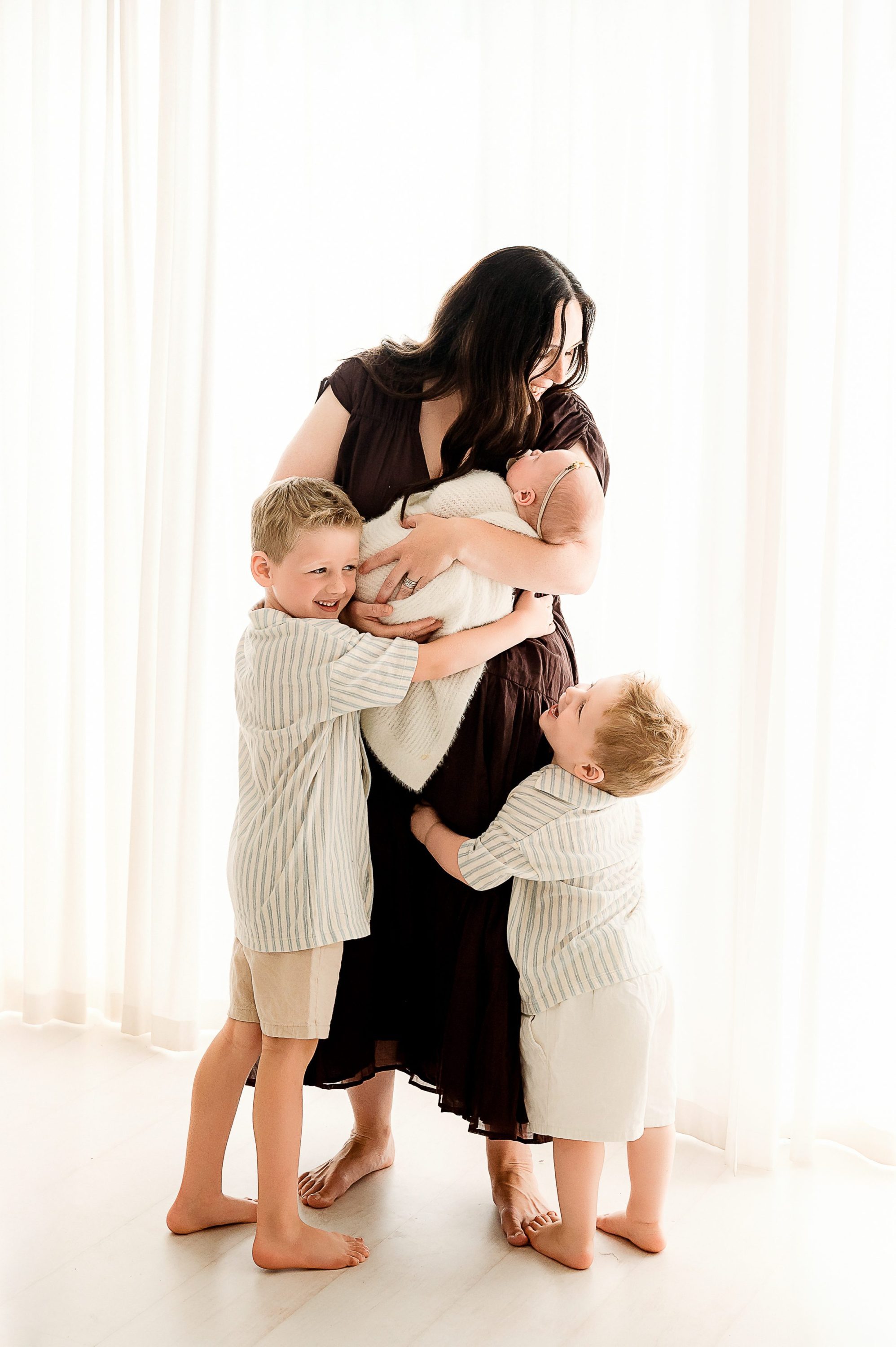 A woman stands in front of sheer white curtains, cradling her baby in a heartfelt Newborn Photography Melbourne west session. Two young boys hug her legs, all smiling and barefoot, capturing a warm, loving family moment.
